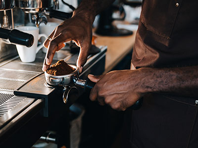 A barista tamps ground wholesale coffee Johannesburg into a portafilter at an espresso machine with a cup in the background. Top Dose Roasters