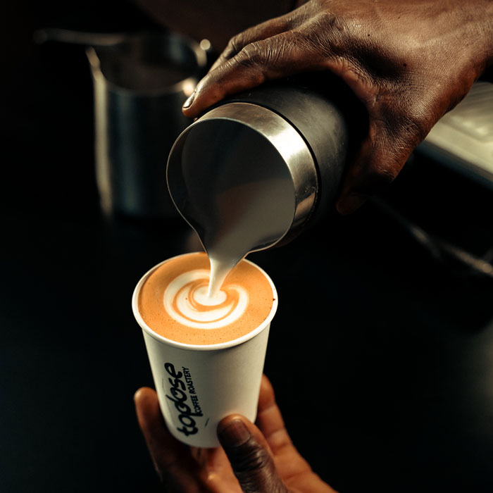 A person pours steamed milk from a metal pitcher, creating latte art on a cup of coffee labeled "Good Coffee," capturing the essence of memorable coffee experiences Johannesburg has to offer. Top Dose Roasters