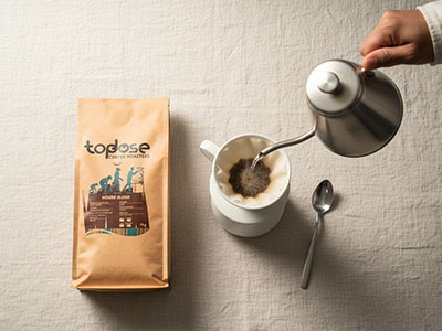 A hand pours hot water from a kettle over wholesale coffee grounds in a dripper above a mug, next to a bag of Topdose wholesale coffee Johannesburg and a spoon on a light-colored tablecloth. Top Dose Roasters