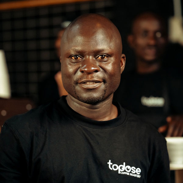 Japhet Matimbe â€“ Head Roaster wearing a black "topdose coffee roastery" t-shirt stands indoors, looking at the camera. Another person in similar attire is visible in the blurred background. Top Dose Roasters