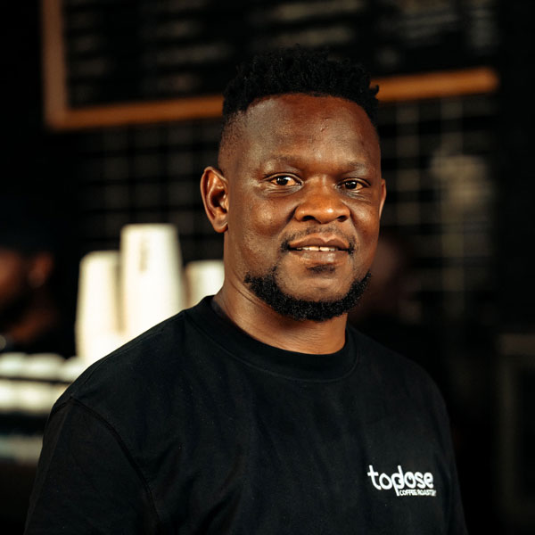 Neo Mkhethoni â€“ Founder & Head Barista wearing a black "topdose" t-shirt stands indoors, with coffee cups and a menu board visible in the blurred background. Top Dose Roasters